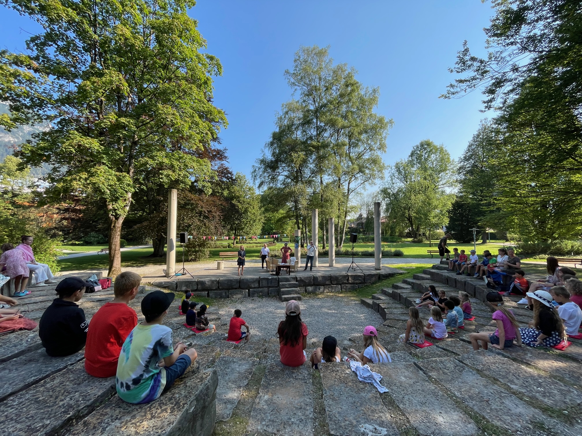 Schülerinnen sitzen im Amphitheater in Garmisch-Partenkirchen. Der Himmel ist blau. Es ist Sommer.