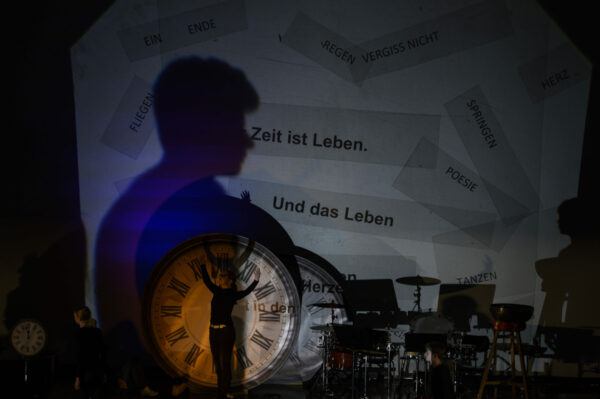 Tänzer steht auf der Bühne mit offenen Armen vor einer Uhr. Im Hintergrund sind verschiedene Sprüche auf der Leinwand erkennbar.